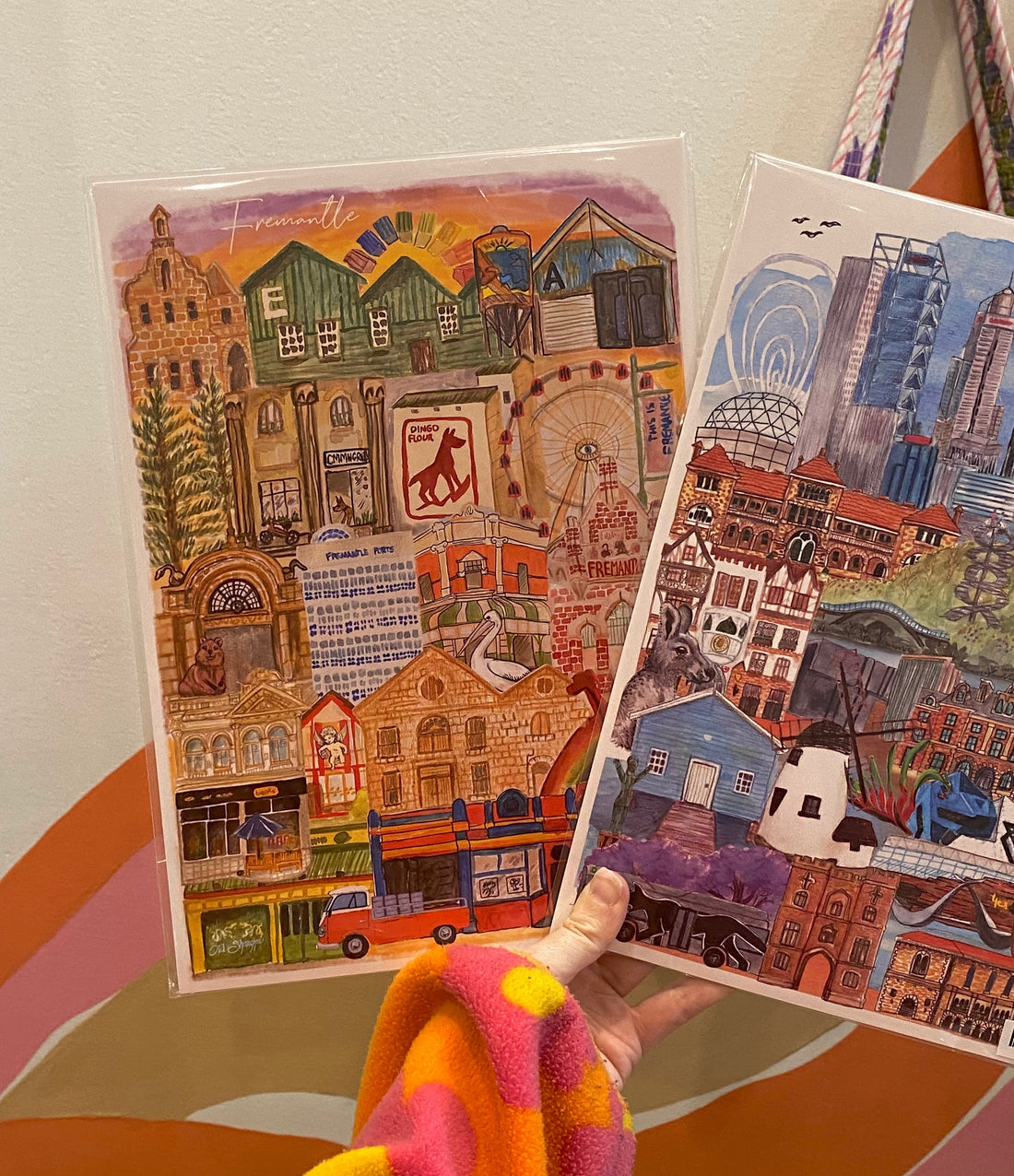 a hand holding two art prints. one of Fremantle and one of Perth