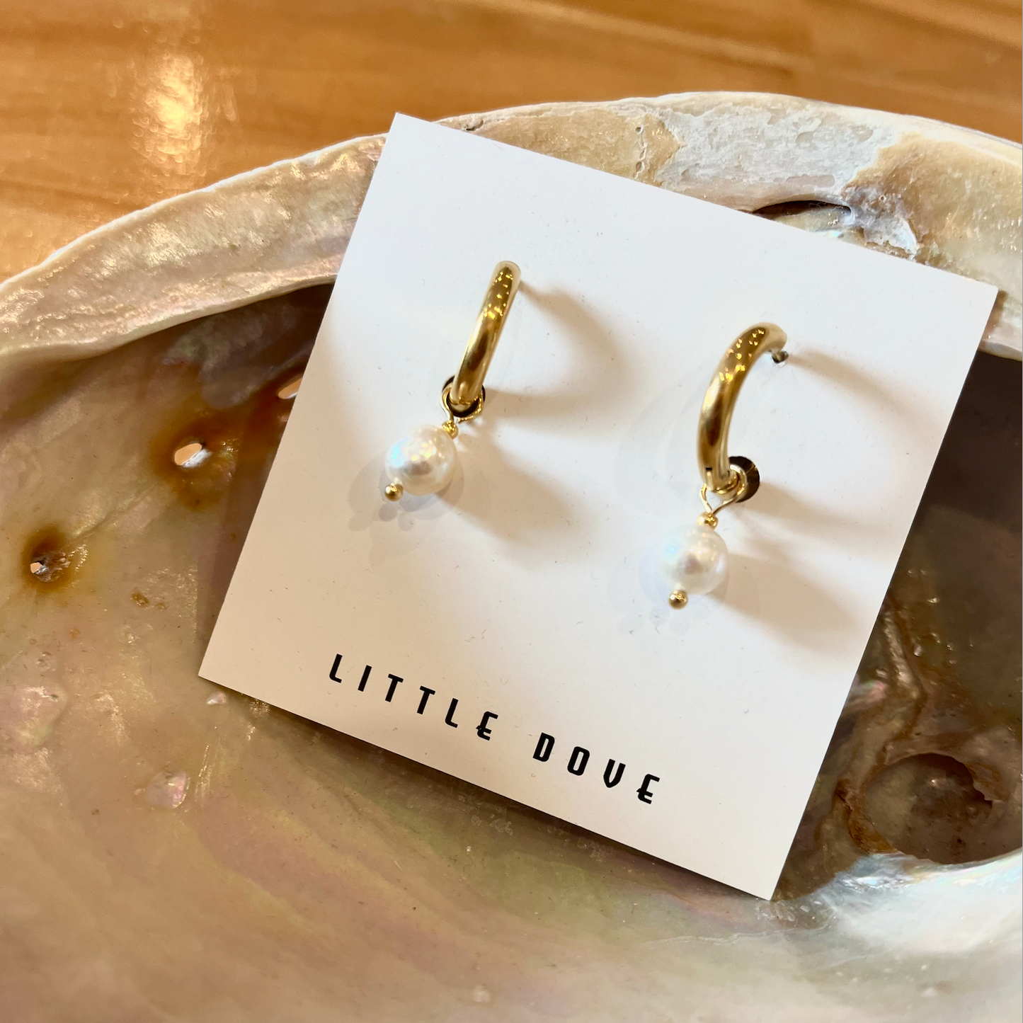 Gold hoop earrings with pearls on a 'Little Dove' branded card, set against a natural stone background.